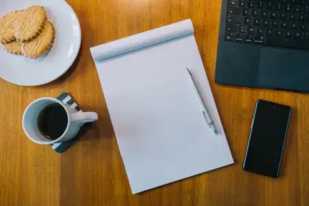 Un bureau avec son bloc note, son ordinateur, son téléphone, sa tasse de café accompagnée de ses gâteaux